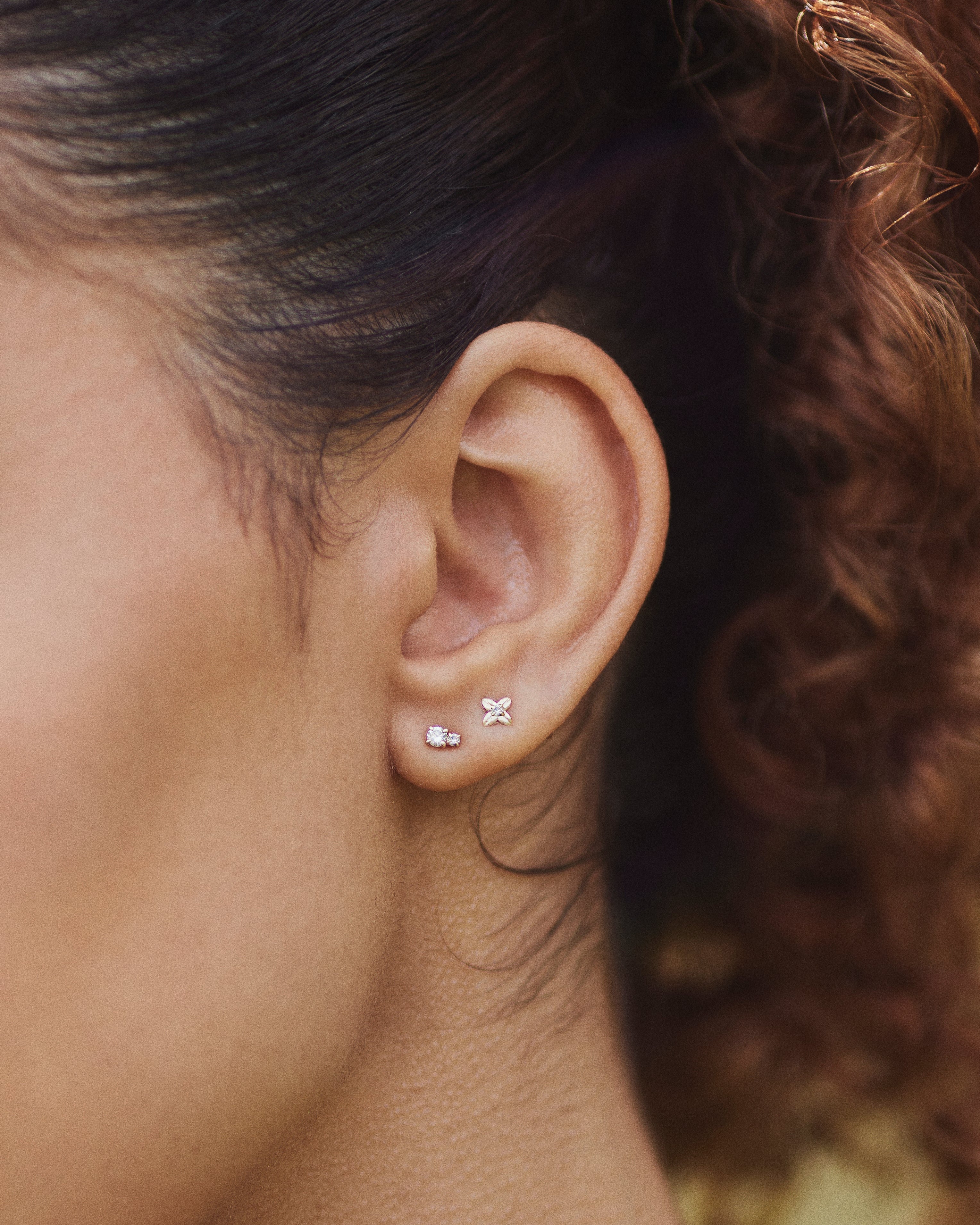 Close-up of a single Flor stud earring shaped like a four-petal flower with a small diamond in the center, shown stacked with our Diamond Duo Stud on the ear.